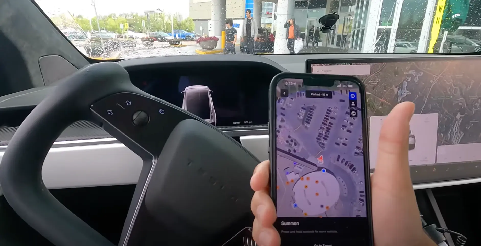 hand holding a smartphone inside a Tesla, using the Smart Summon feature with a map displayed, while the steering wheel and center screen are visible, showing a rainy parking lot outside