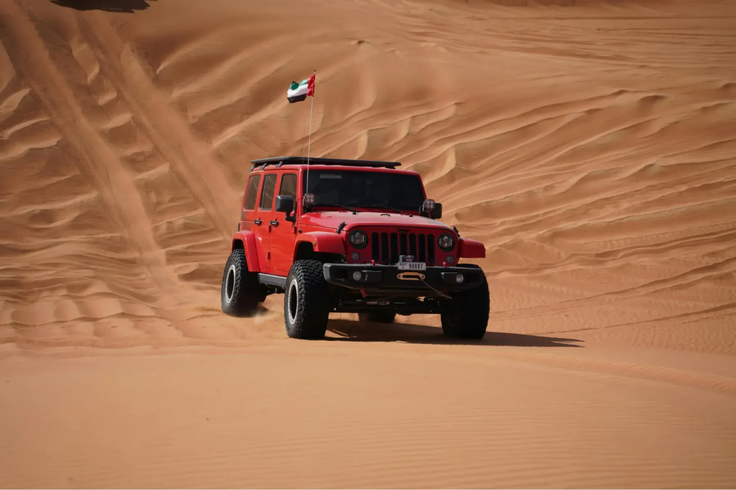 Jeep Wrangler suspension upgrades - JC Whitney Jeep Wrangler mods featured on a red Wrangler with a roof rack, off-road tires, and a dune flag, driving through sandy terrain.