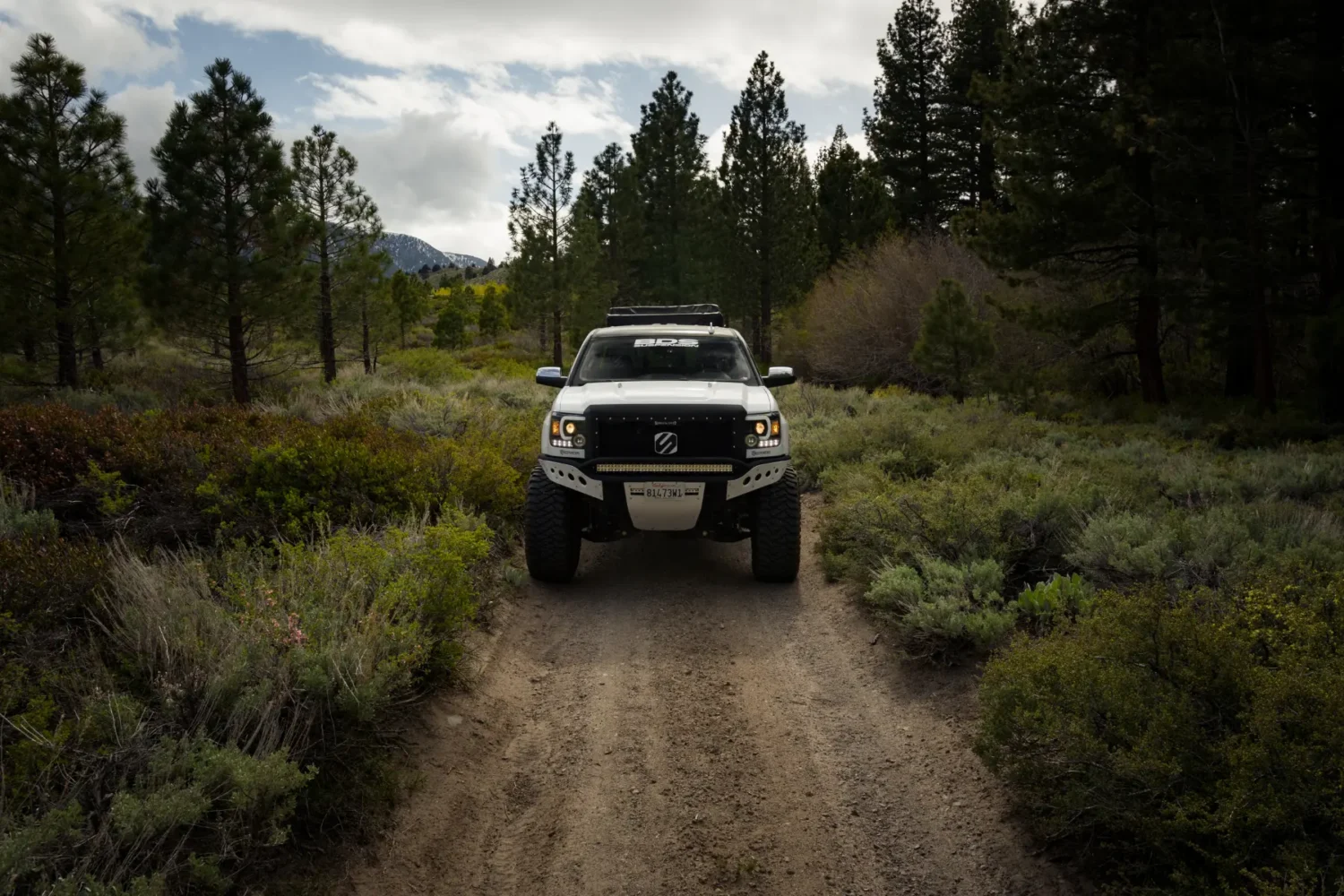 off-road suspension kits - JC Whitney Off-road suspension kits enhance a white lifted truck navigating a dirt road through a dense forest with mountains in the background.